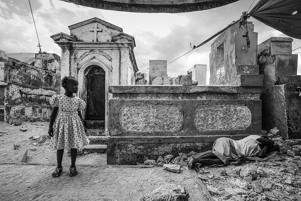 'MOURNING IN THE CEMETERY' - PORT-AU-PRINCE, HAITI. JANUARY 12, 2015. On the fifth anniversary of the devastating earthquake, a young child stands to the side of her mourning mother who lies weeping nearby in the Grand Cemetery of Port-Au-Prince, Haiti.